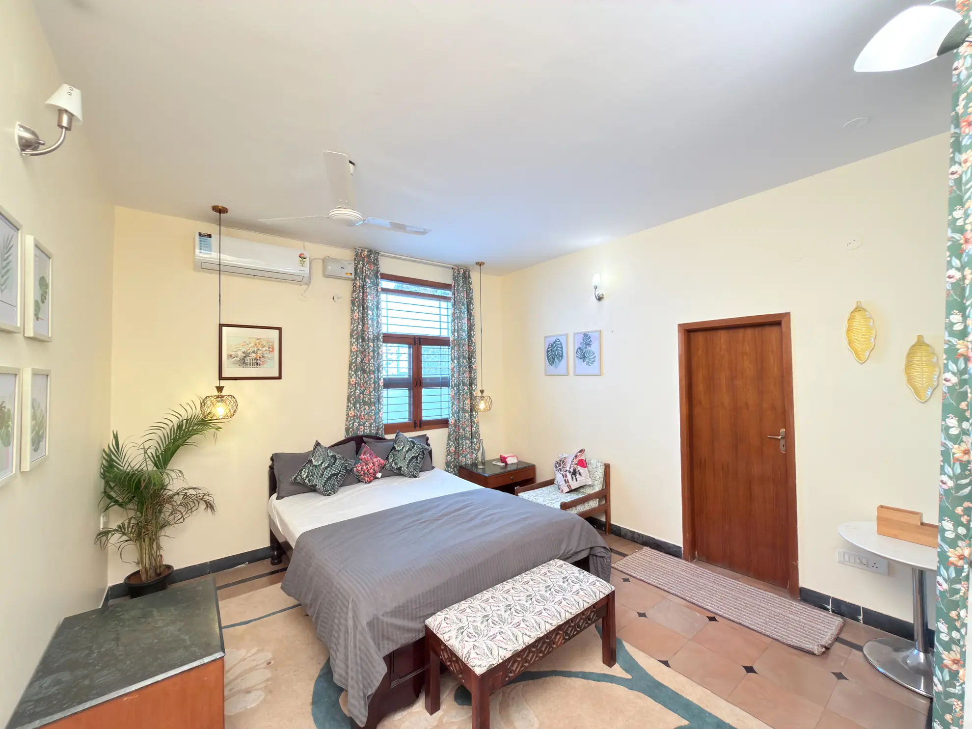 Bedroom with a queen bed, wall-mounted air conditioner, and a potted palm beside patterned curtains and a window.