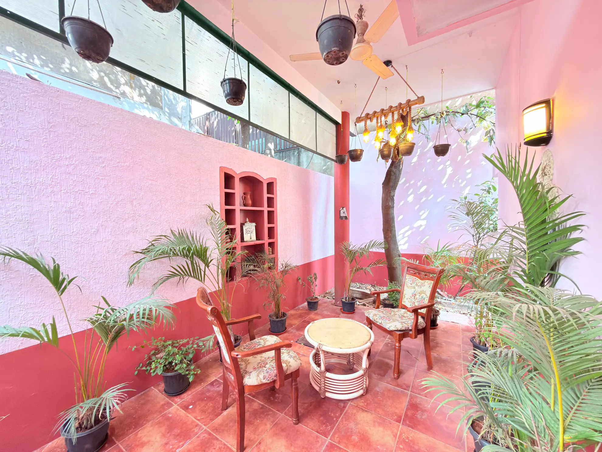 Pink-walled covered patio with two wooden armchairs, a round table, hanging planters and tall potted palms.