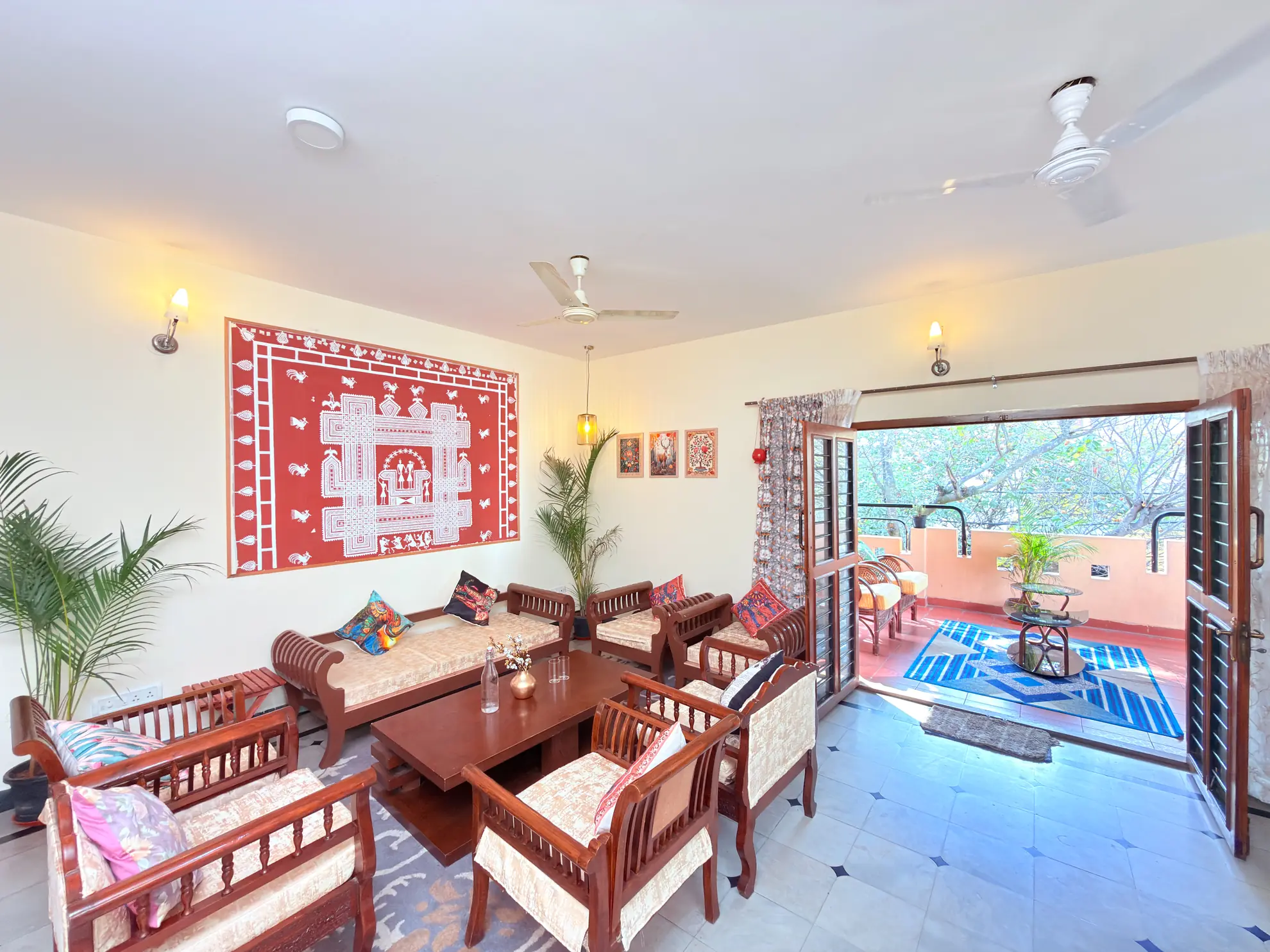 Living room with wooden sofa set, red wall hanging, and open doors onto a sunlit terrace with plants.