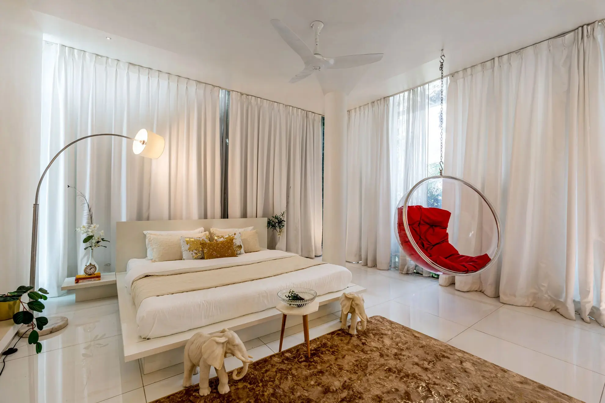 Bright white bedroom with a king bed, red hanging bubble chair by floor-to-ceiling curtains, and elephant figurines.