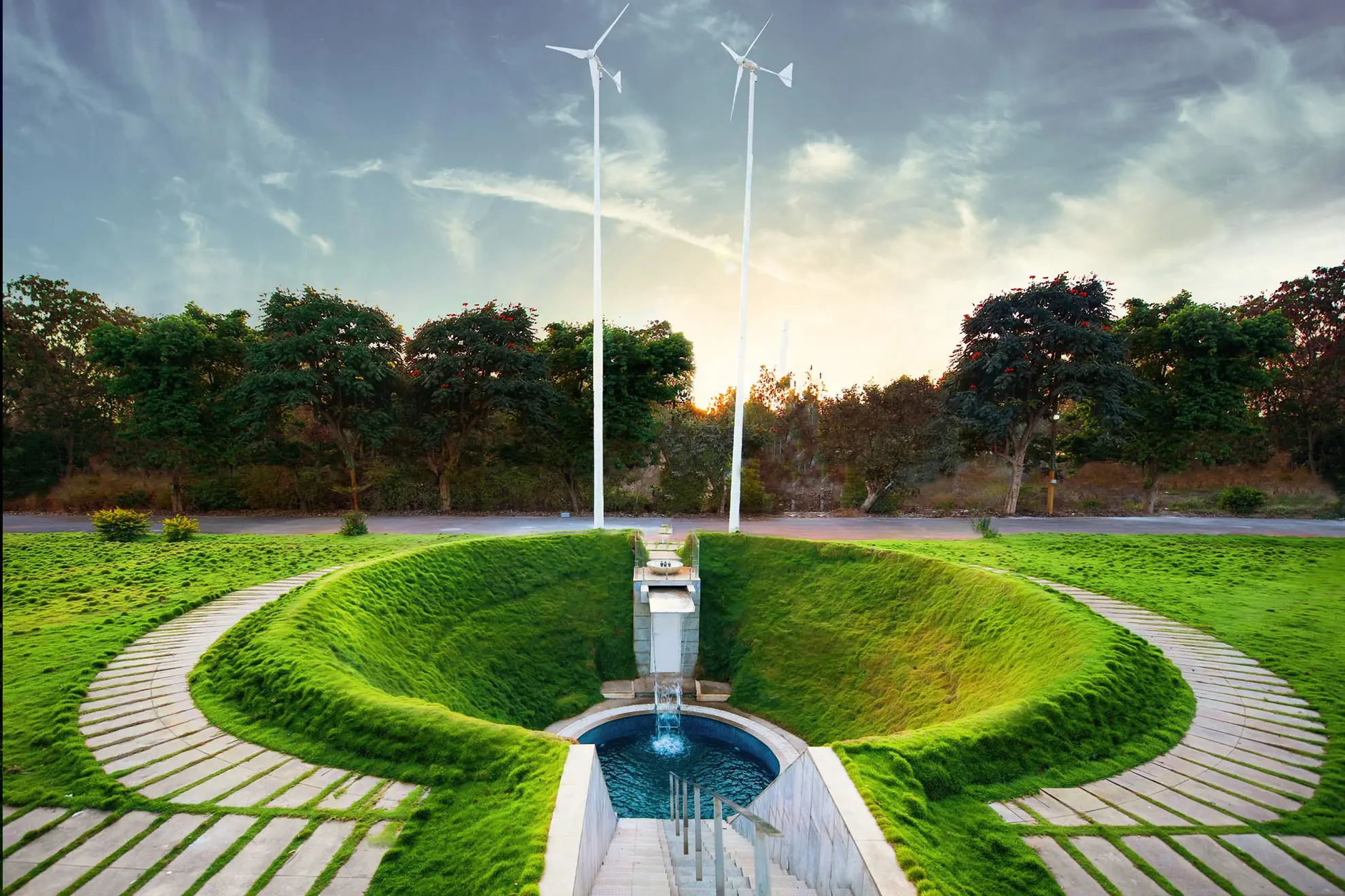 Grass-lined sunken pool with a small fountain at its base, wind turbines and trees behind it.