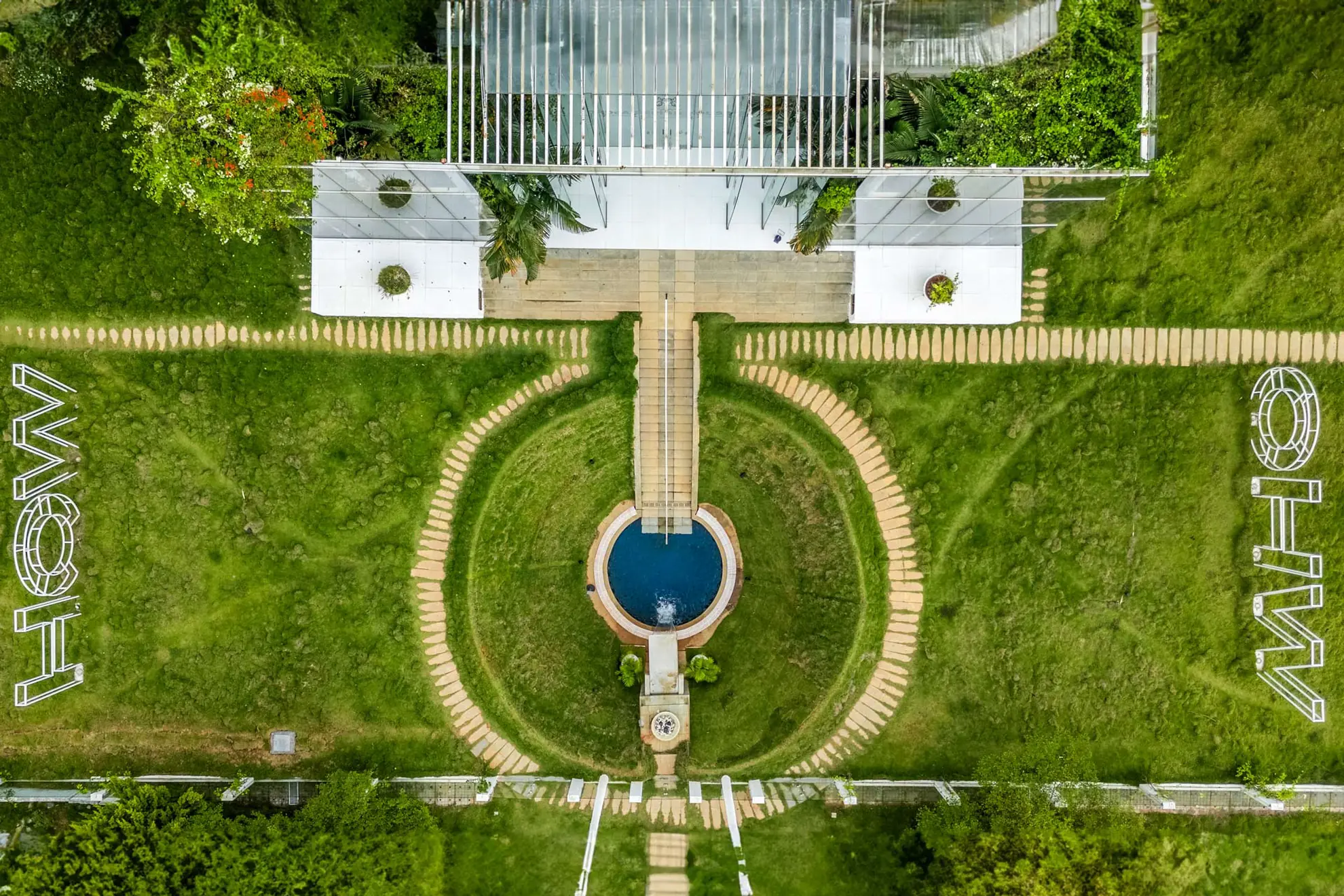 Overhead view of a round blue pool at the end of a long central path, flanked by lawns and a glass-roofed pavilion.