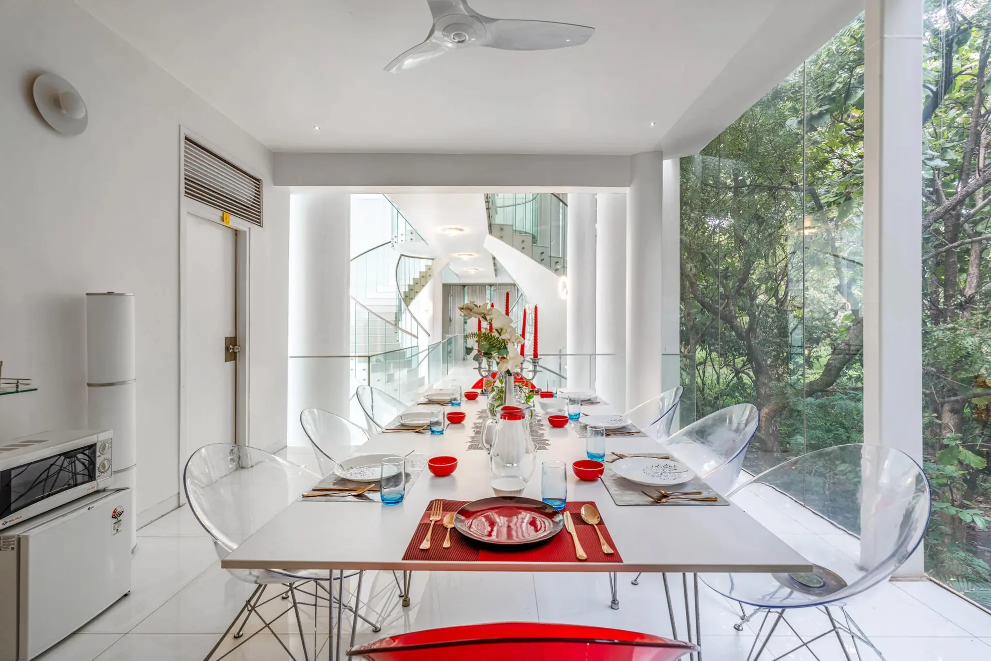 White dining room with a long table set in red, glass walls onto lush trees, and a spiral staircase beyond.
