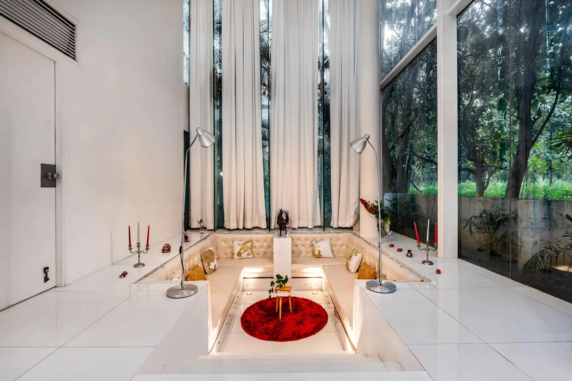 Sunken white living area with tufted banquette seating, a red round rug, and floor-to-ceiling windows onto trees.