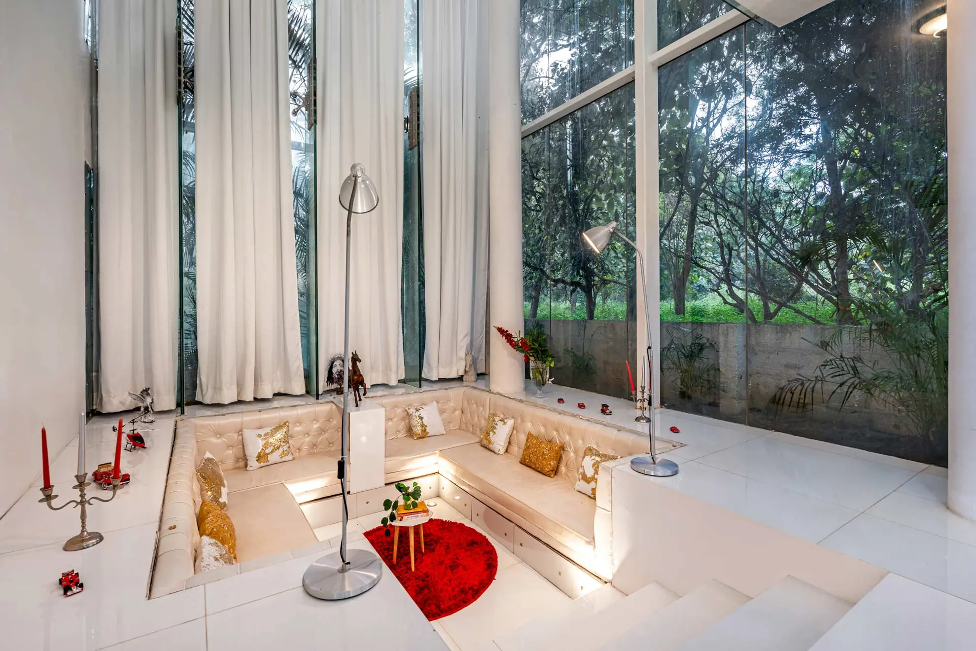 Sunken white lounge with tufted banquette and gold cushions, beside floor-to-ceiling glass overlooking greenery.