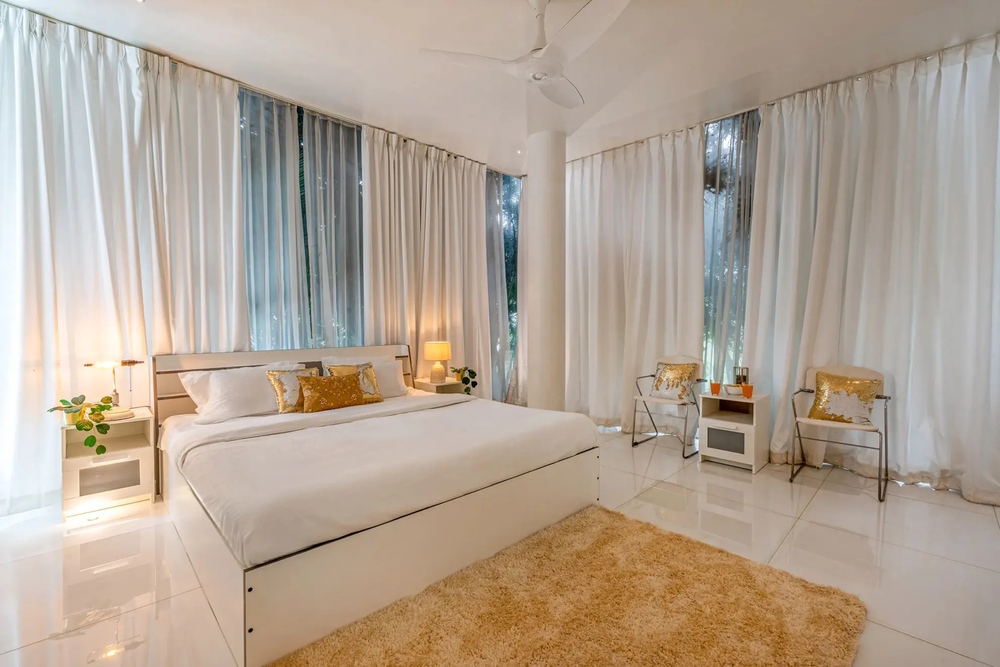 Bright white bedroom with a king bed, gold cushions, sheer curtains and a ceiling fan.