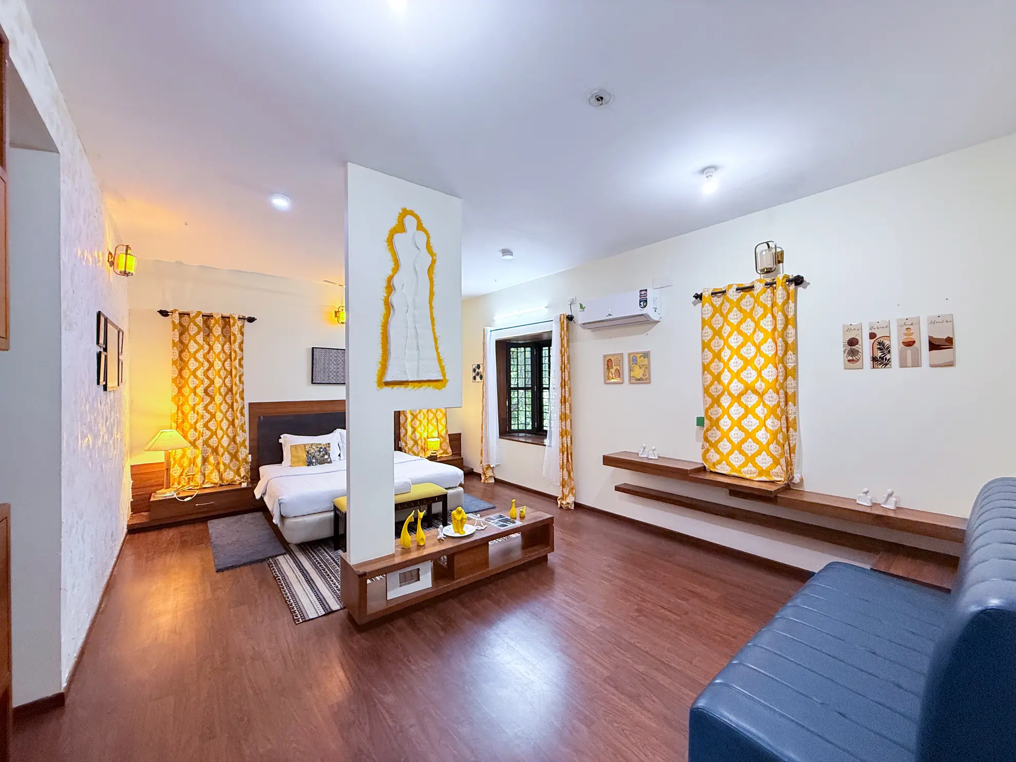 Open-plan room with a double bed, yellow patterned curtains, a wall-mounted air conditioner and a blue leather sofa.