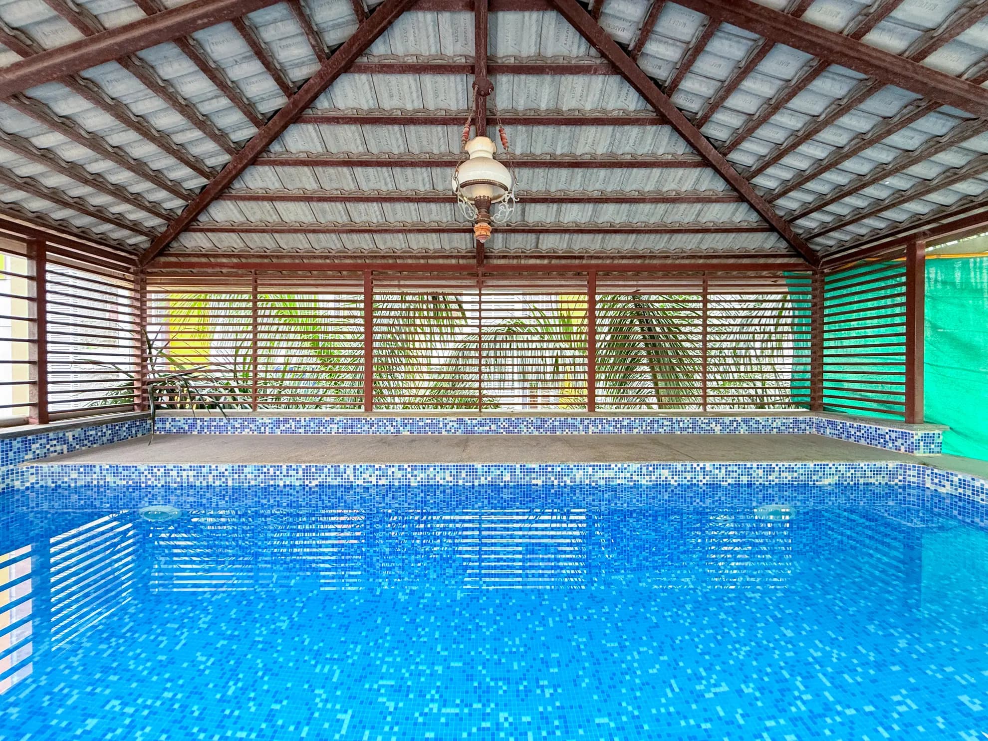 Indoor swimming pool with blue mosaic tiles under a wooden-beamed roof, with a hanging lantern.