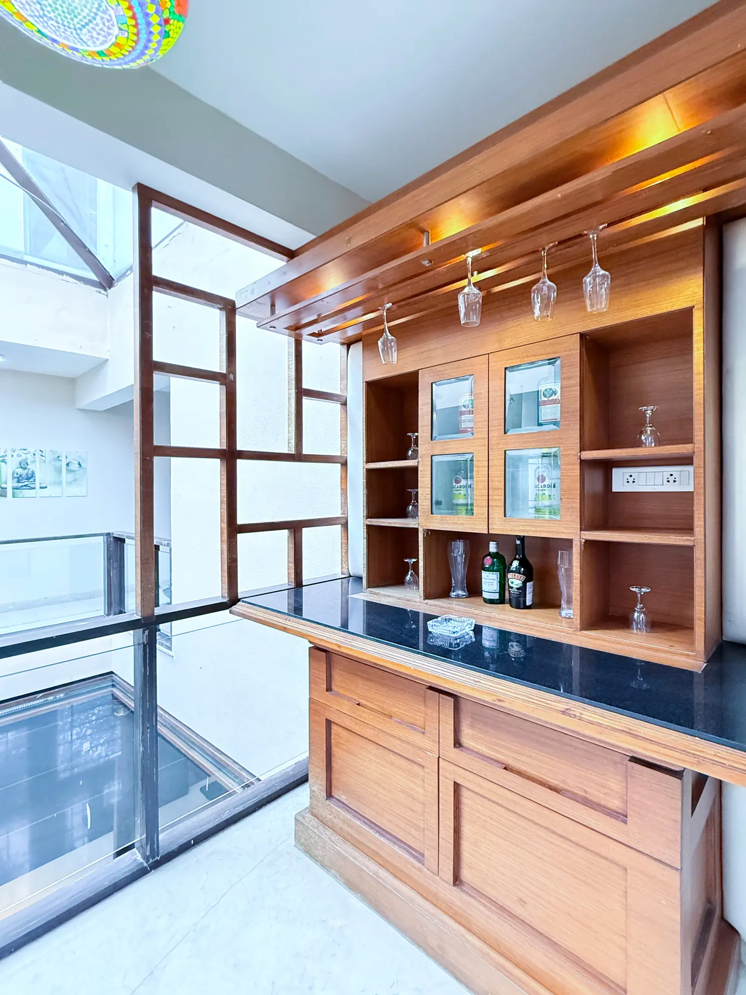 Built-in wooden bar with glass-front cabinets, hanging stemware, and a black stone counter beside a glass wall.