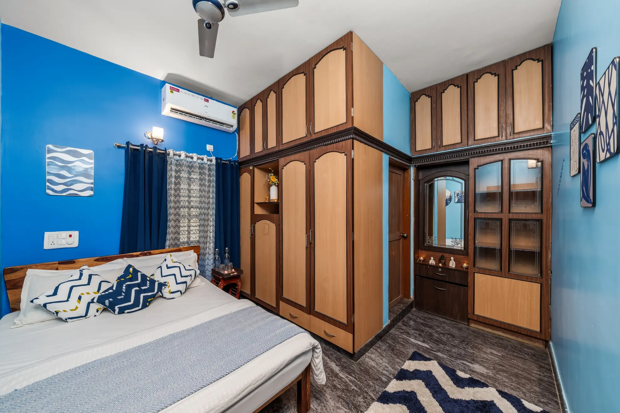 Blue-walled bedroom with a double bed, tall wooden wardrobes, a wall-mounted air conditioner and a ceiling fan.
