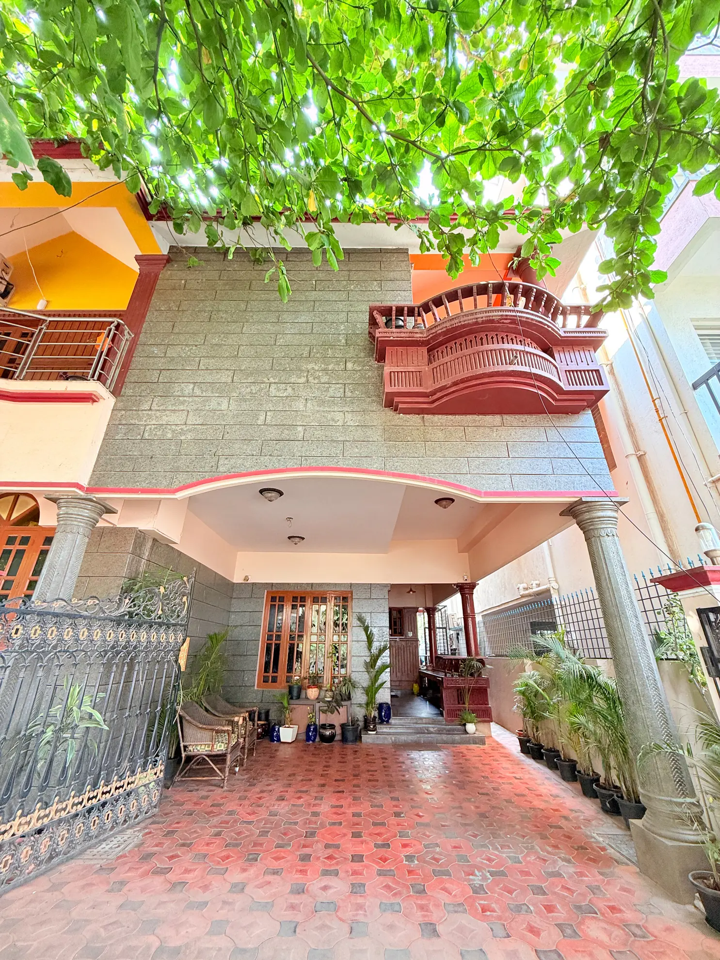 Front of a two-storey house with a red-tiled forecourt, potted plants, a carved red balcony and a wrought-iron gate.