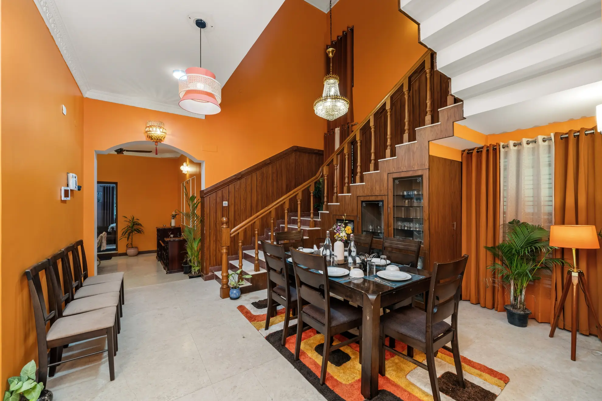 Dining area with a dark wood table set for six under a chandelier, beside a wood-paneled staircase and bright orange walls.