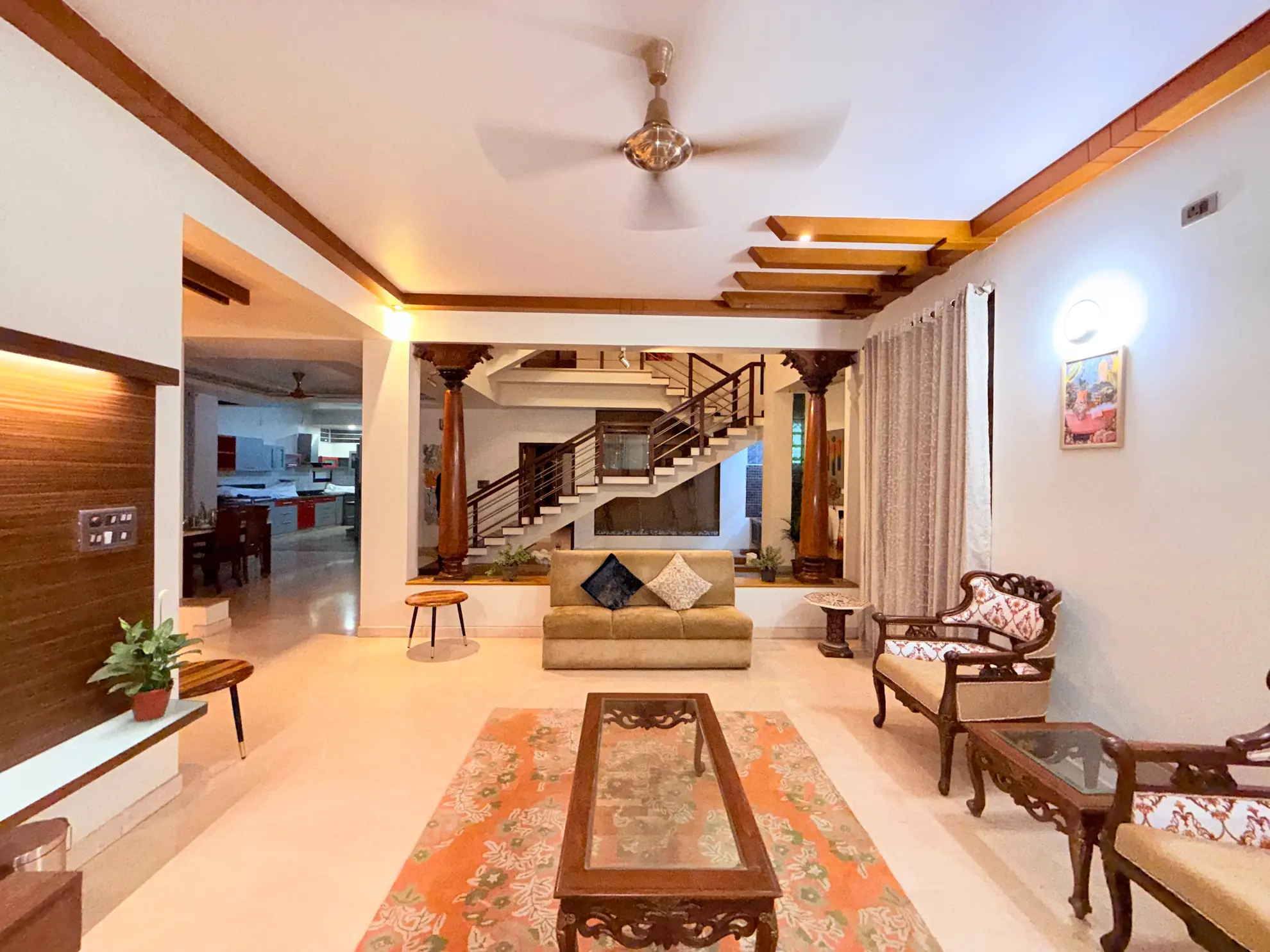 Open living room with patterned rug, wooden coffee table, and timber staircase rising behind a tan sofa.