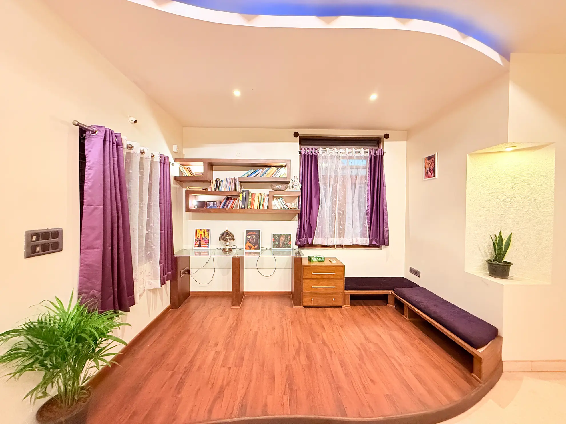 Bright room with a glass-top desk, floating wooden bookshelves, and a cushioned bench seat by the window.