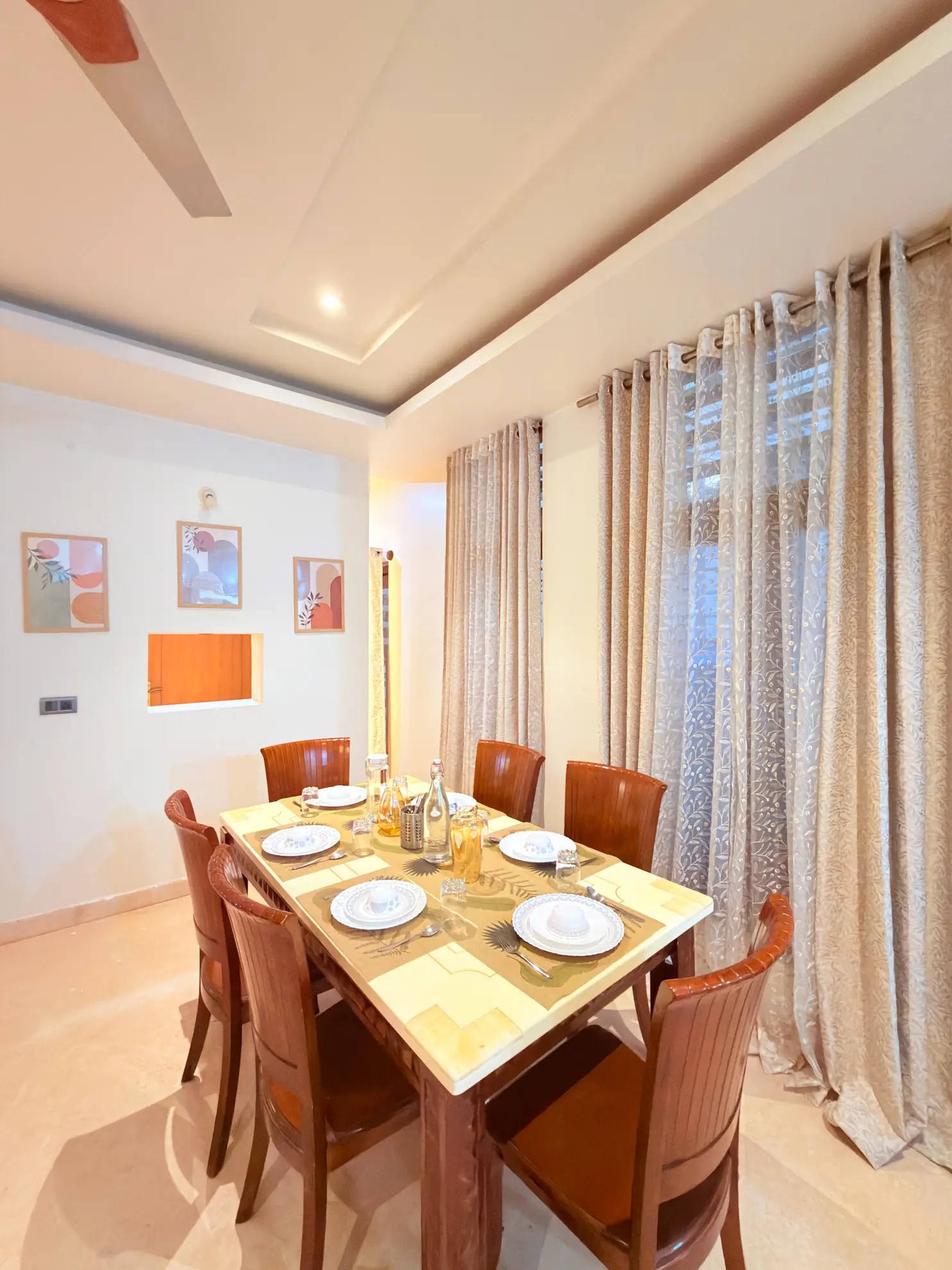 Dining room with a six-seat wooden table set for a meal, framed wall art, and sheer patterned curtains by the window.