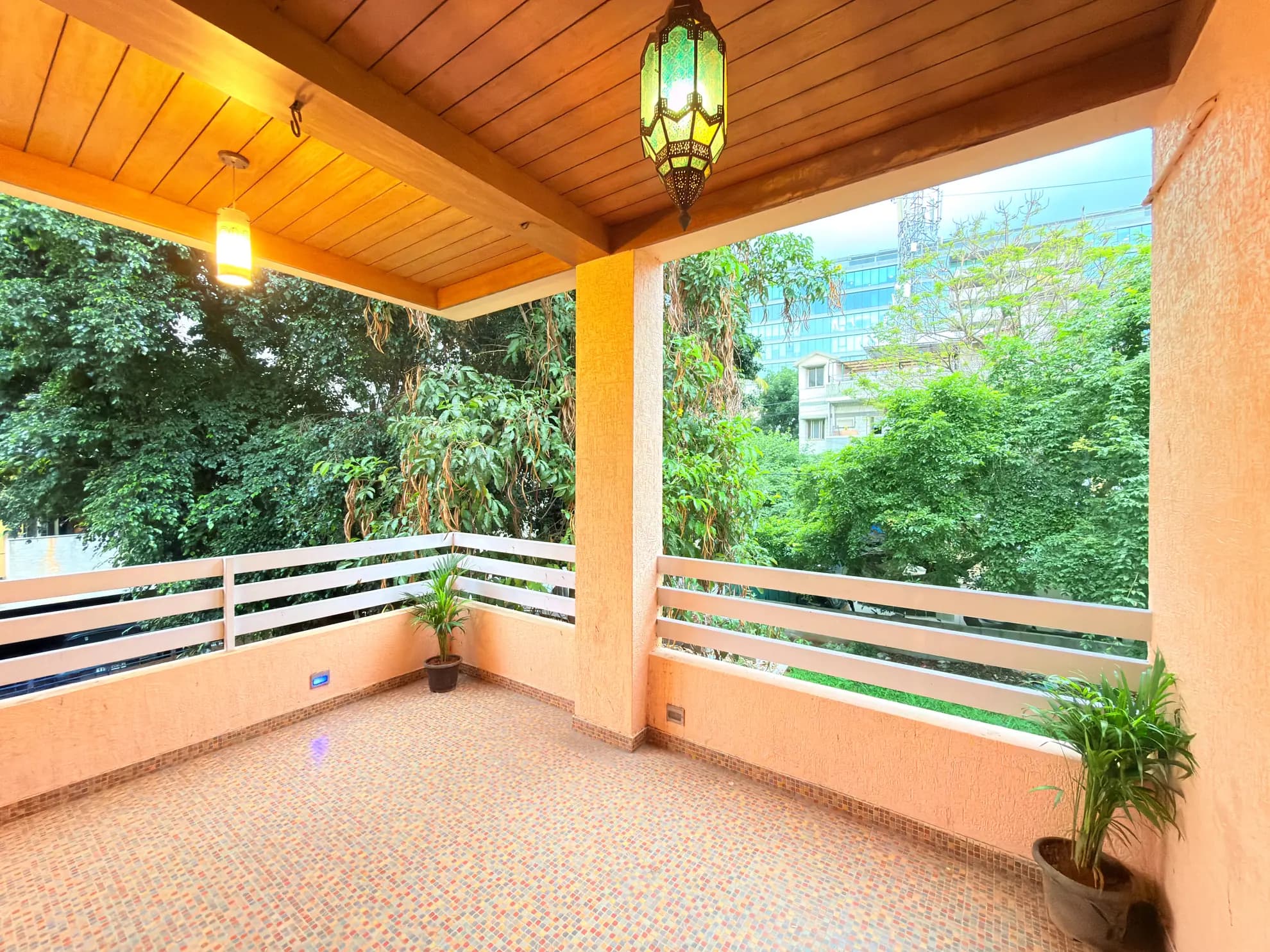Covered balcony with tiled floor, glass lantern, and white railings overlooking green trees and a building.