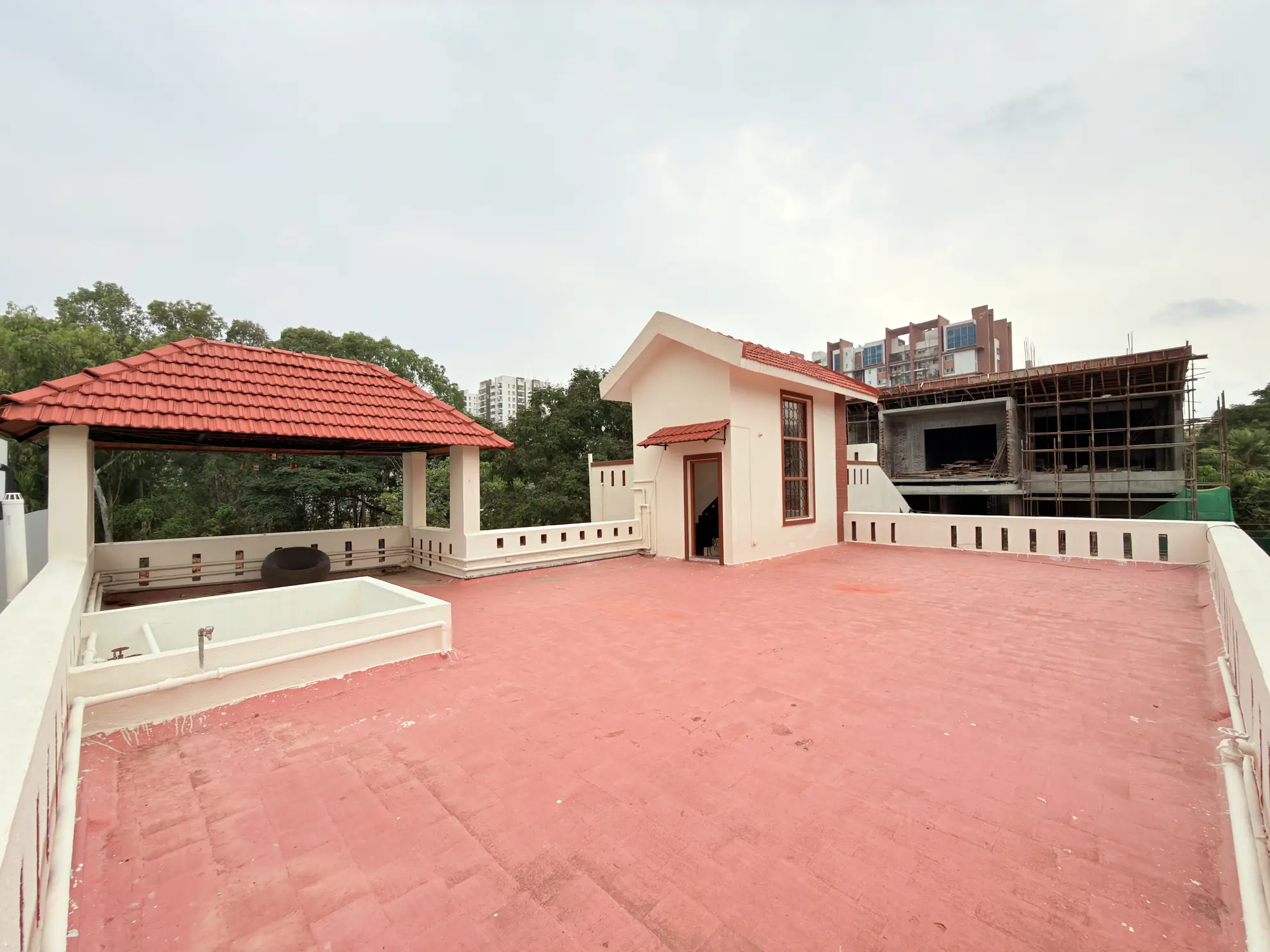Open rooftop terrace with red-tiled covered pavilion, small cream utility building and an empty tub, with a construction site beyond.