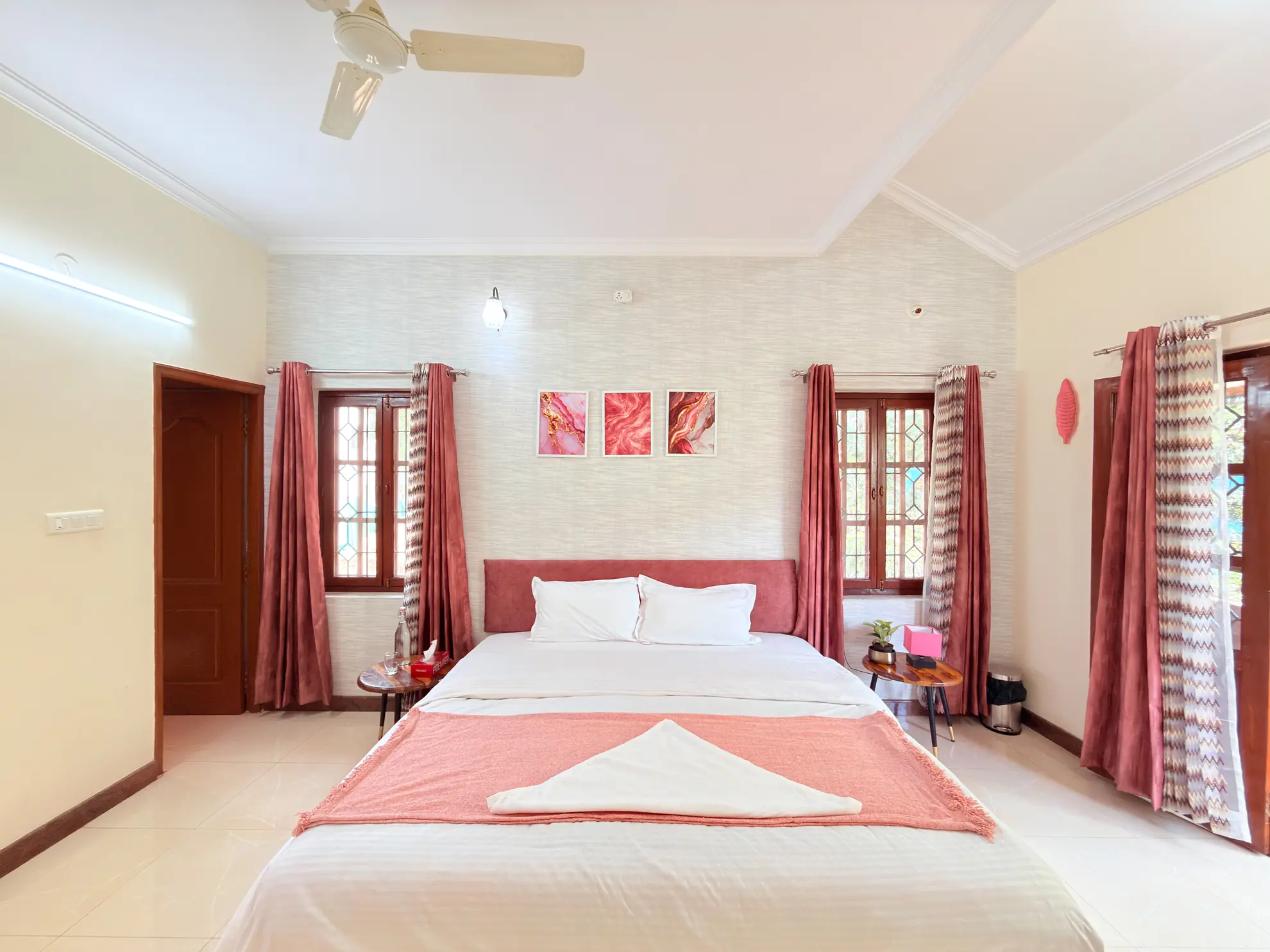 Bedroom with a made queen bed, pink accent cushions and curtains, and a ceiling fan overhead.