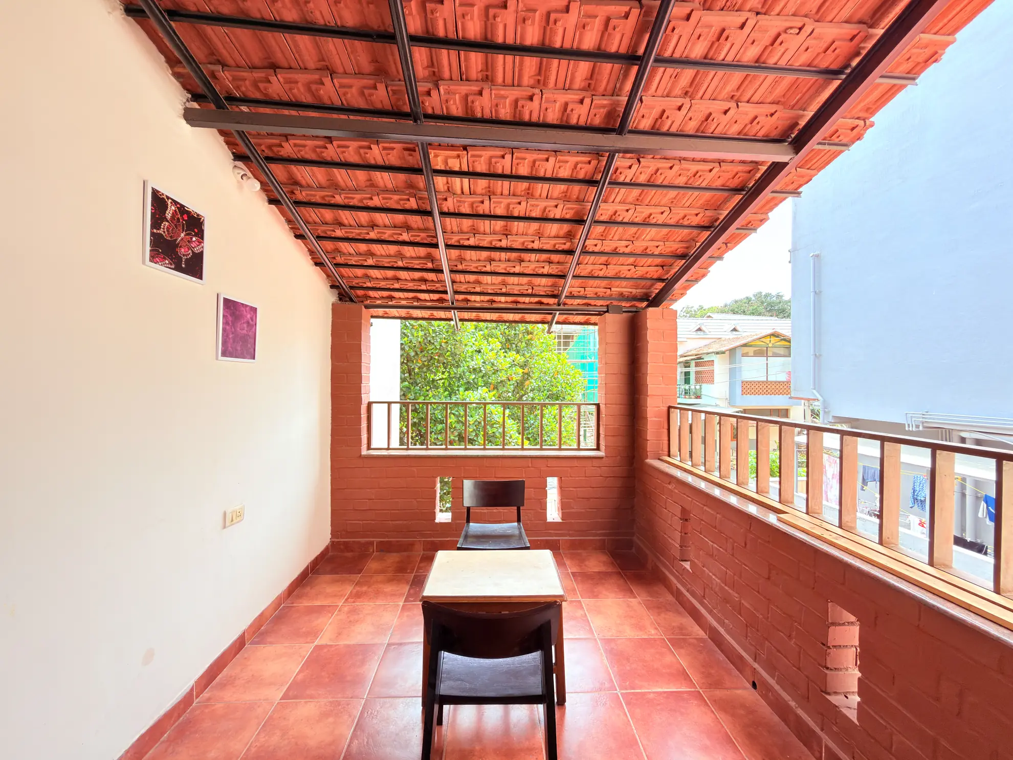 Covered terracotta-tiled balcony with a small wooden table and chair, a leafy view and a railing.