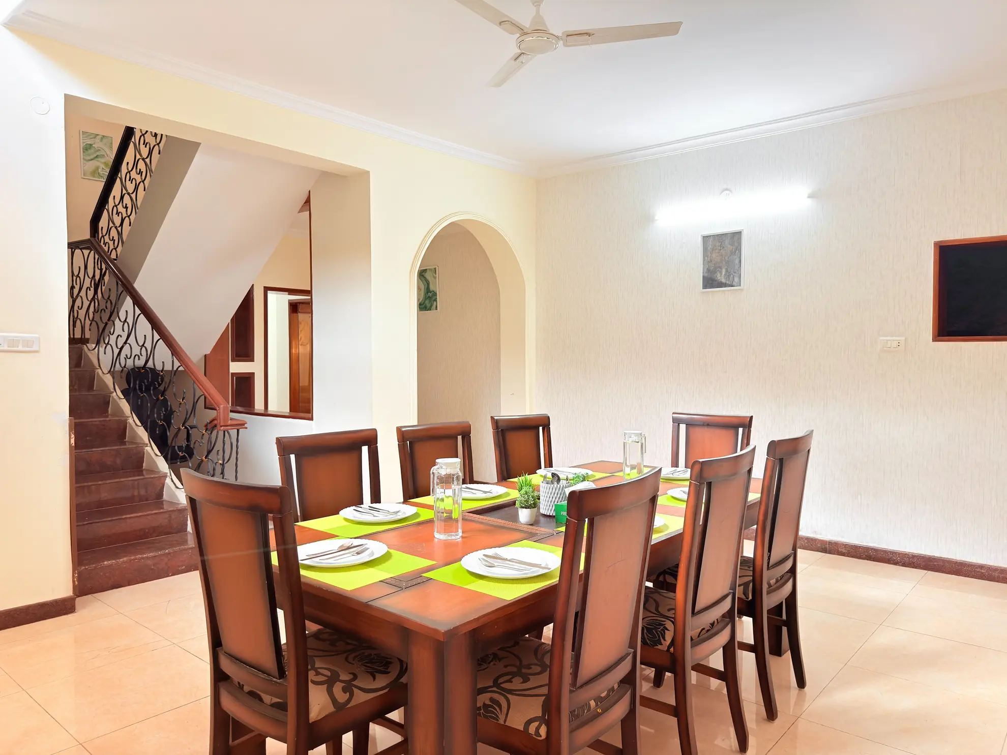 Dining area with a wooden table set for eight, green placemats, and a staircase with ornate iron railing.