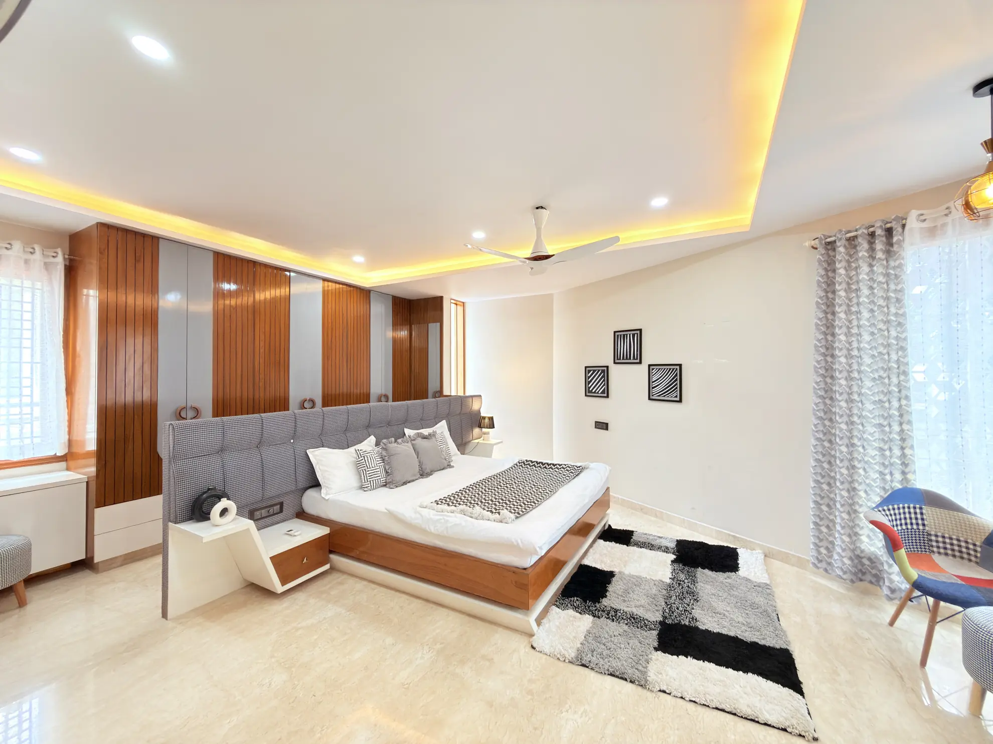 Bedroom with a large upholstered bed, warm backlit ceiling cove, and a patchwork armchair by the window.