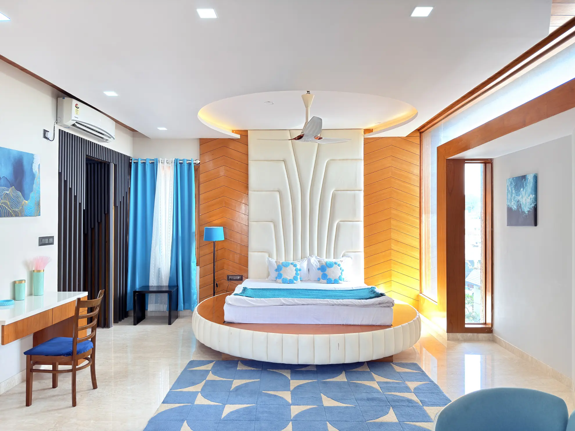 Bedroom with a round raised bed set on a blue patterned rug, wooden wall panels, and a sheer blue curtain.