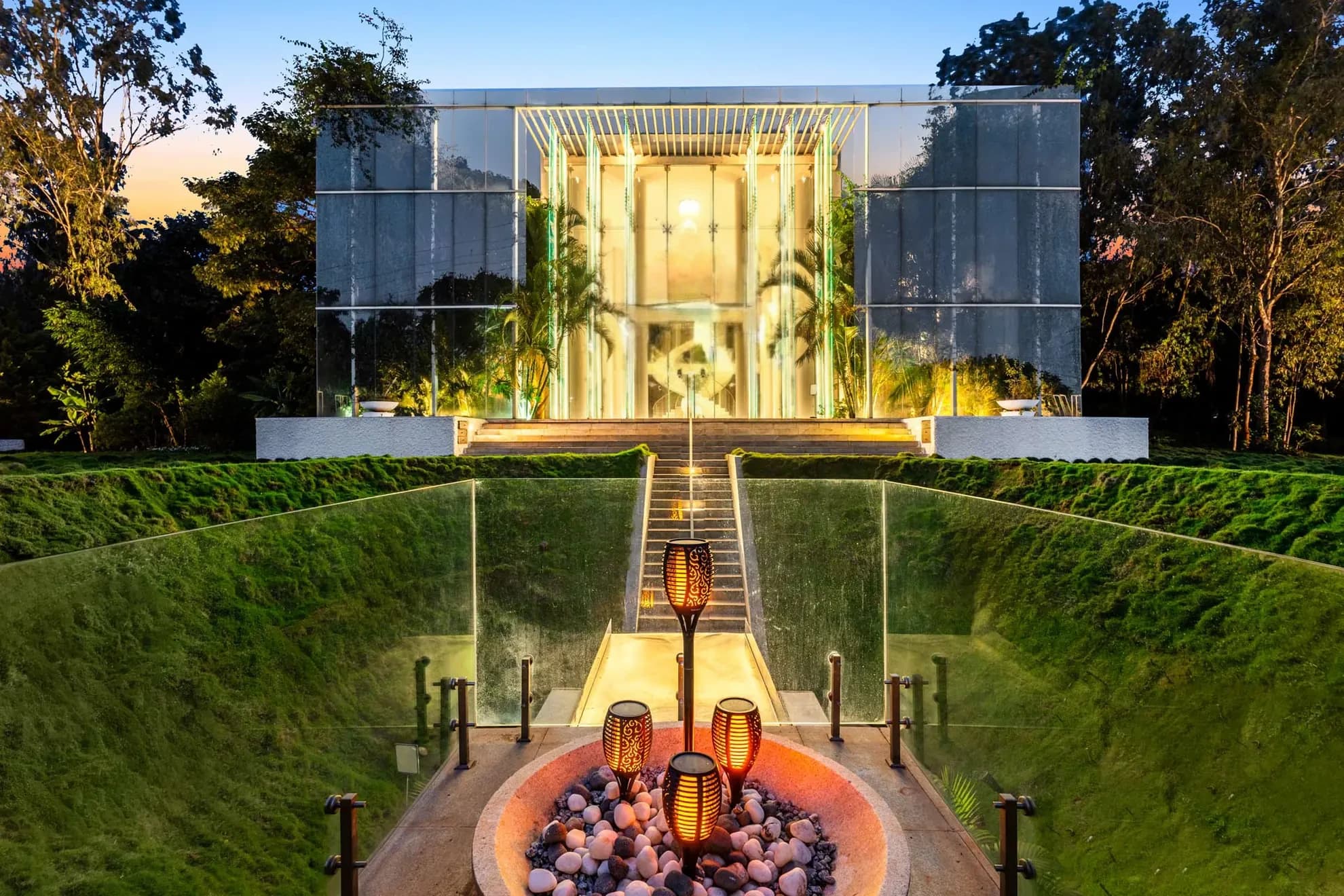 Crystal Hall at dusk — a glass-walled villa lit from within, above a sunken courtyard with a lit fire bowl