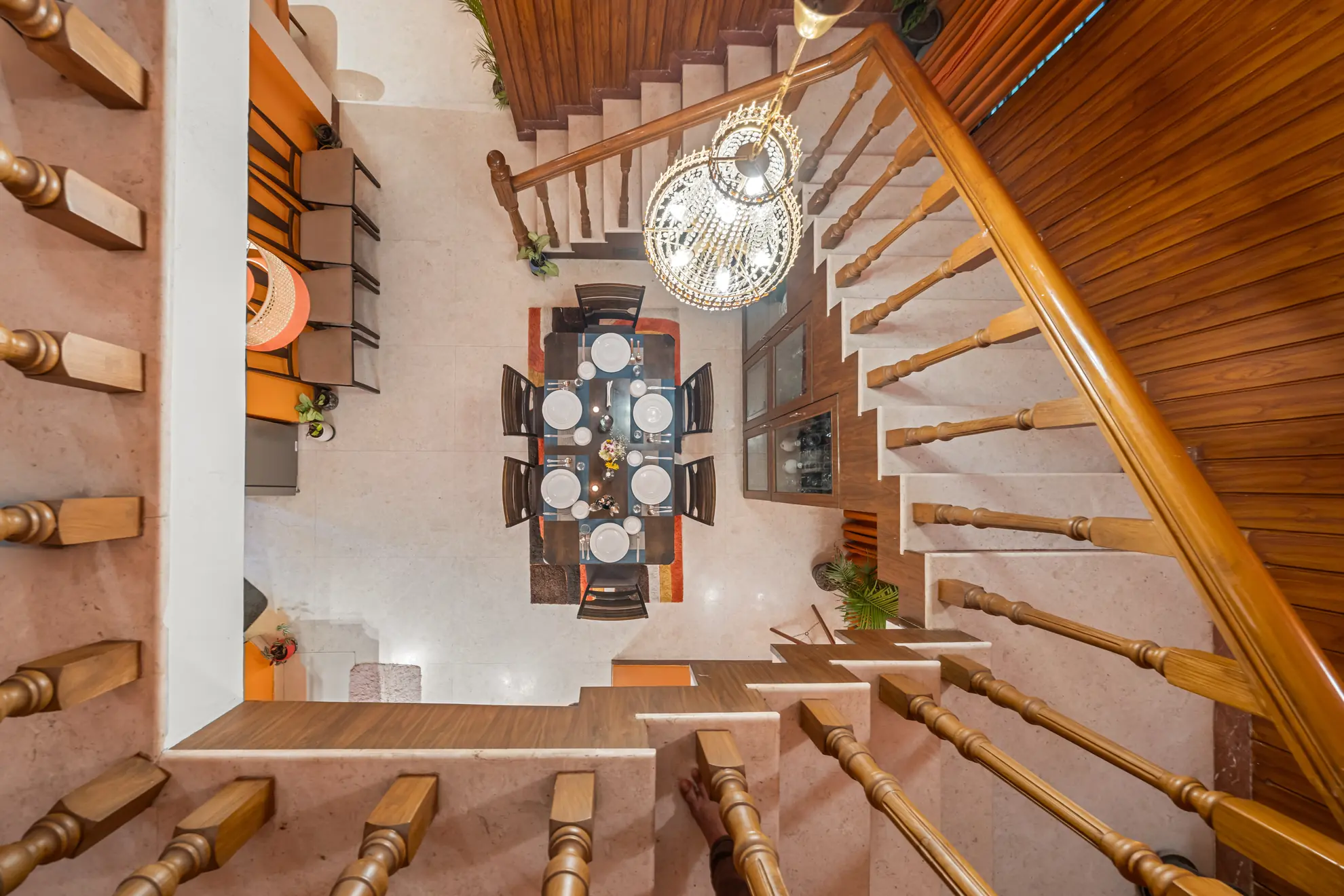Overhead view of a dining table set for four beneath a crystal chandelier, beside a wooden staircase with turned balusters.